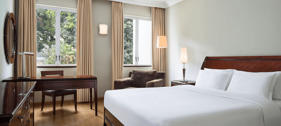 Modern and cozy hotel room with a large white bed, wooden furniture, soft lighting, beige curtains, and large windows overlooking lush greenery.