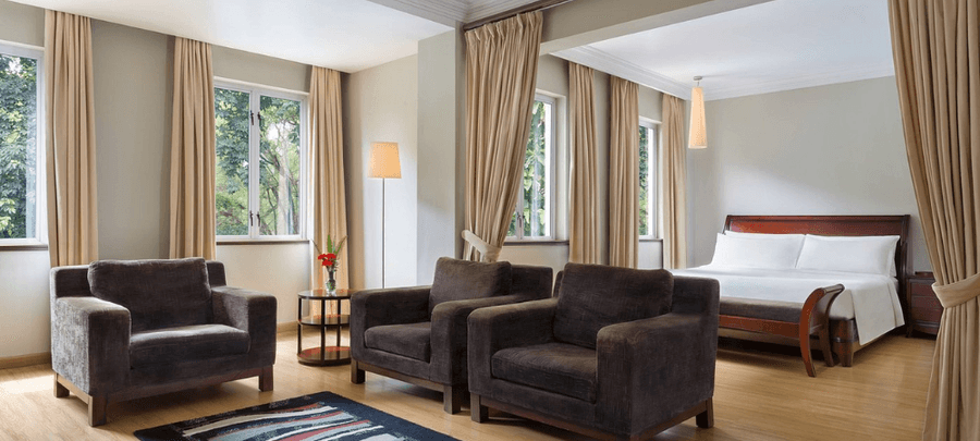 Bright and spacious hotel suite featuring a large bed with white linens, three plush armchairs, tall windows with beige curtains, wooden flooring, and soft natural lighting overlo…