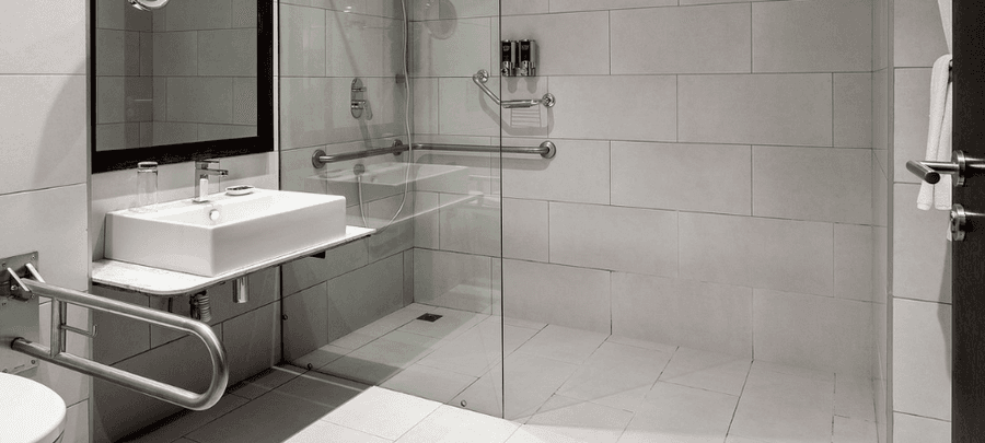 Modern accessible bathroom with a walk-in glass shower, grab rails, tiled walls, and a sleek white sink.