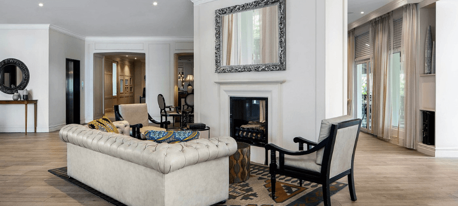 Elegant hotel lounge with modern décor featuring a cream tufted sofa, armchairs, a fireplace with a decorative mirror, large windows with sheer curtains, and spacious wooden floor…