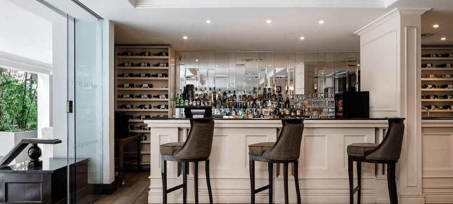 Stylish modern hotel bar with high stools, a mirrored back wall lined with liquor bottles, built-in wine shelves, and soft ambient lighting.