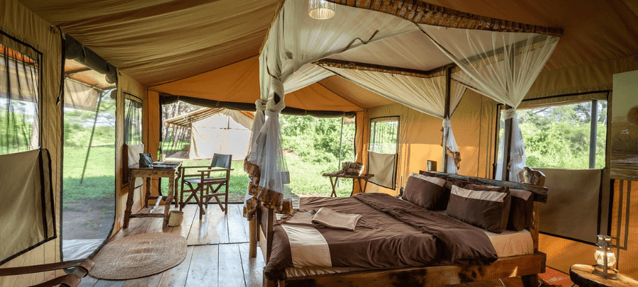 A spacious safari tent bedroom with a wooden four-poster bed draped in mosquito netting and dressed in earthy brown linens. Large screened windows and open canvas walls allow natu…