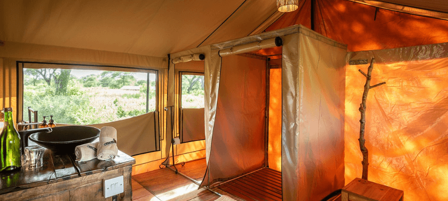 A bright and rustic safari tent bathroom area featuring a freestanding basin sink on a wooden vanity, rolled white towels, and a private enclosed shower space. Large screened wind…