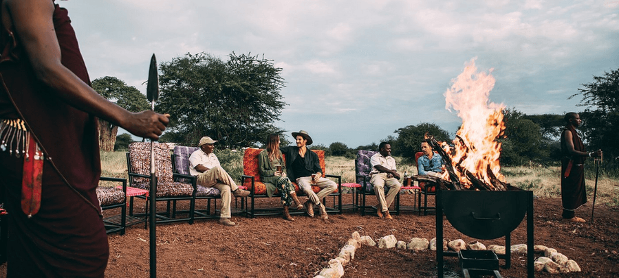 Guests and guides gather around a large campfire in an open safari setting, enjoying conversation and storytelling as Maasai warriors stand nearby in traditional attire.