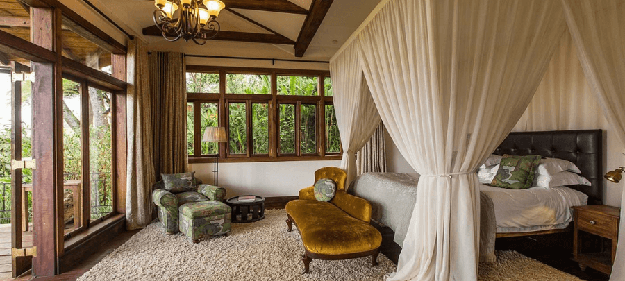 Elegant bedroom suite with a four-poster canopy bed draped in white fabric, cozy seating area, large wooden-framed windows, and warm rustic décor overlooking lush greenery.