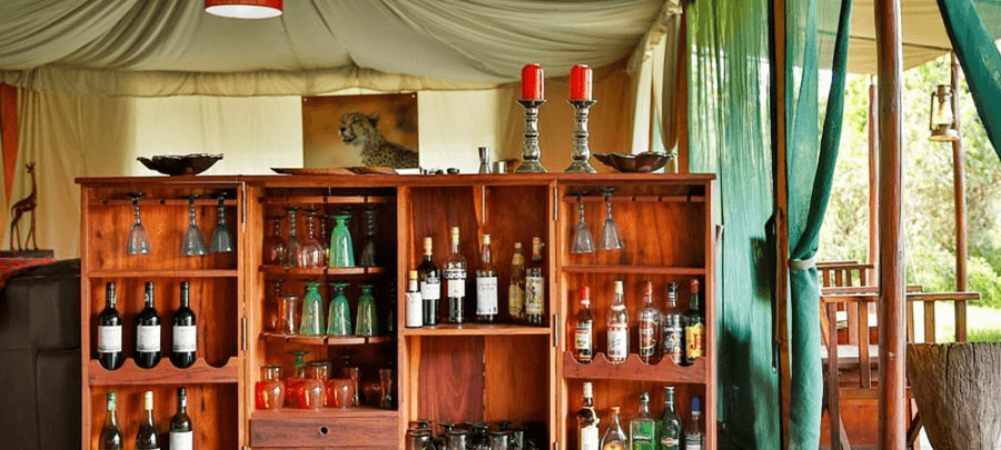 A rustic wooden bar cabinet stocked with wine bottles, spirits, and colorful glassware sits inside an elegant safari tent lounge. Red candles, draped canvas ceilings, and open gre…