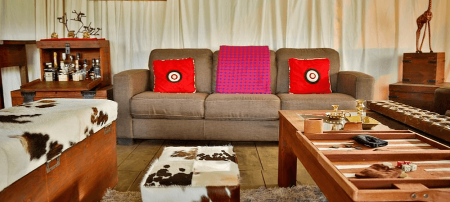 A cozy safari-style lounge area featuring a neutral-toned sofa with bright red accent cushions, surrounded by warm wooden furniture and cowhide-covered ottomans. A stocked minibar…