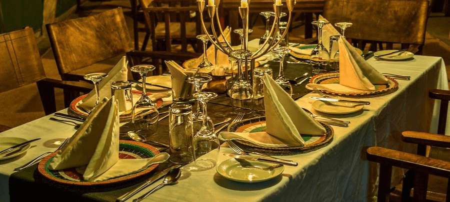 An elegant dining table set for dinner with neatly folded white napkins, polished glassware, silverware, and colorful woven placemats. Warm ambient lighting and a decorative candl…