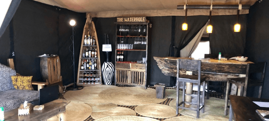Rustic safari lounge and bar area featuring wooden furnishings, warm hanging lights, and a stocked drinks station labeled “The Waterhole.”