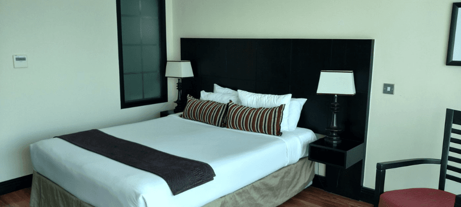 Modern hotel bedroom with a large neatly made bed, striped accent pillows, matching bedside lamps, dark wood furnishings, and polished wooden floors.