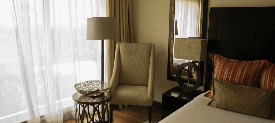 Stylish hotel room corner with a cozy armchair, bedside lamp, decorative table, and neatly made bed beside large sheer-curtained windows letting in soft natural light.