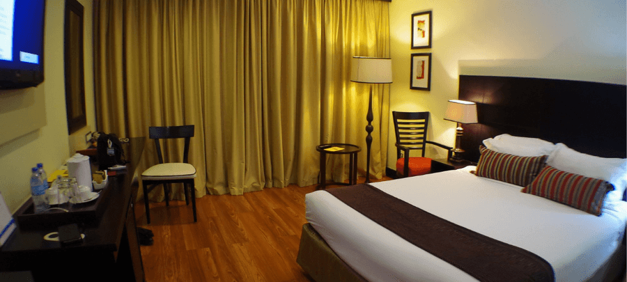 Cozy hotel room with a large bed, warm lighting, wooden floors, bedside lamps, and floor-to-ceiling curtains creating a comfortable atmosphere.