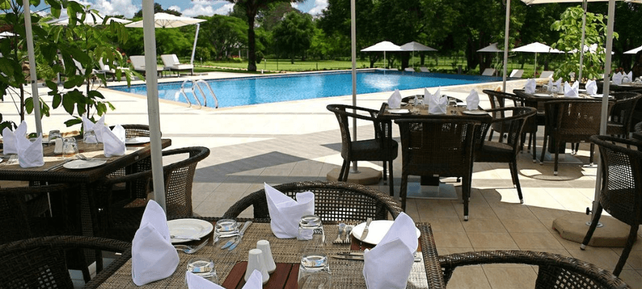 Outdoor poolside dining area with neatly set tables, shaded seating, and a large swimming pool surrounded by lush greenery and lounge chairs.