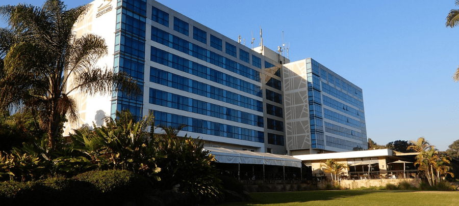 Large modern hotel building with reflective glass windows, surrounded by palm trees, landscaped gardens, and open green spaces under a clear blue sky.
