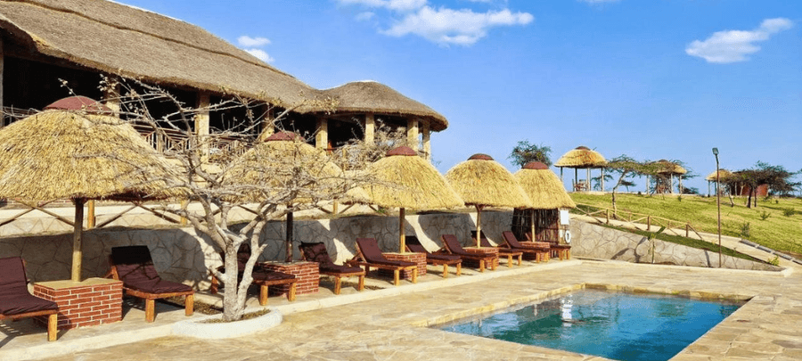 A scenic lodge-style pool area with cushioned sun loungers shaded by thatched umbrellas beside a small swimming pool. In the background, a large thatched-roof building overlooks g…