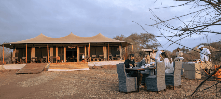 Guests relax around a glowing campfire at dusk outside a luxury safari tent camp, enjoying drinks and conversation under the open evening sky. Warm firelight contrasts with the so…