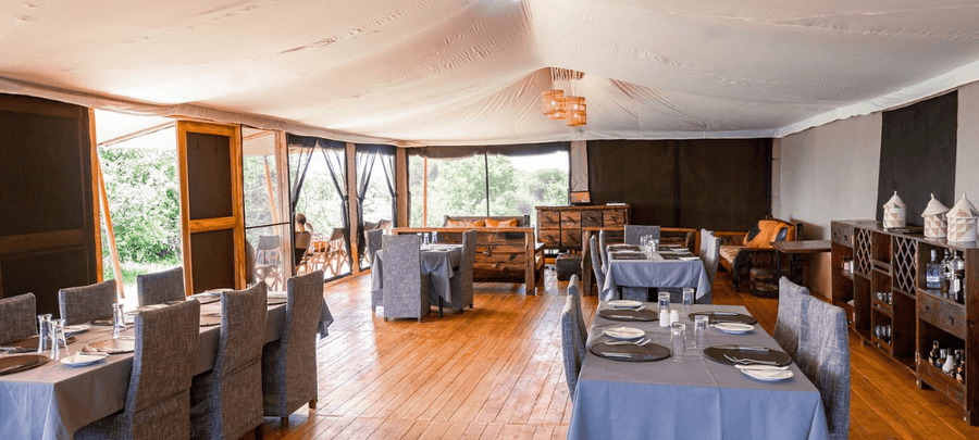 A spacious and elegant safari dining tent featuring polished wooden floors, neatly arranged dining tables, and contemporary rustic décor. Large screened openings allow natural lig…