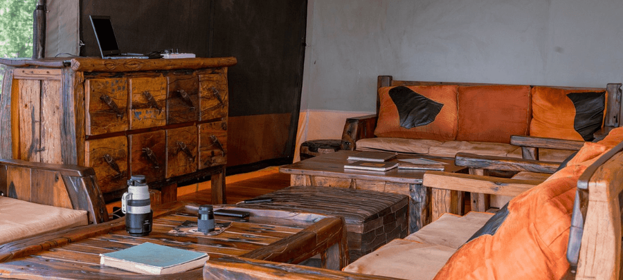A cozy safari lounge area furnished with handcrafted wooden sofas, rustic coffee tables, and warm earth-toned cushions inside a luxury canvas tent. Natural wood textures, books, a…