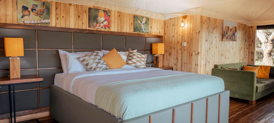 A spacious safari-style bedroom features a large king bed with crisp white linens, patterned accent cushions, and warm bedside lighting. Natural wood-paneled walls, wildlife artwo…