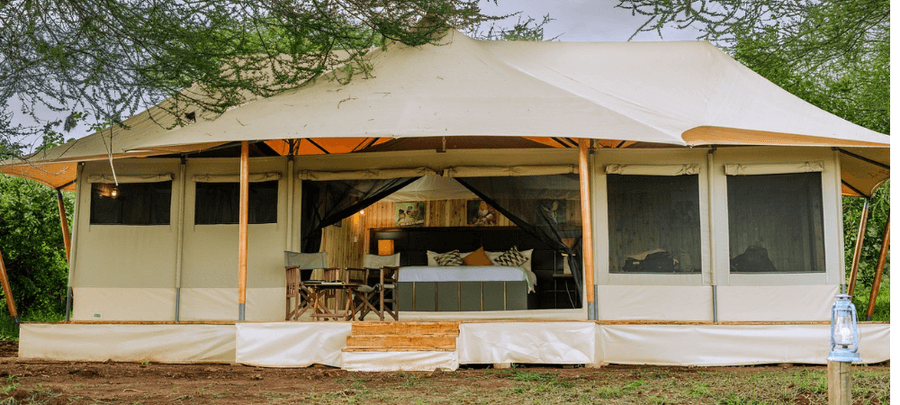 A luxury safari tent sits beneath leafy trees, featuring a spacious canvas structure with screened sides and a wooden deck entrance. Inside, a comfortable bed and seating area are…