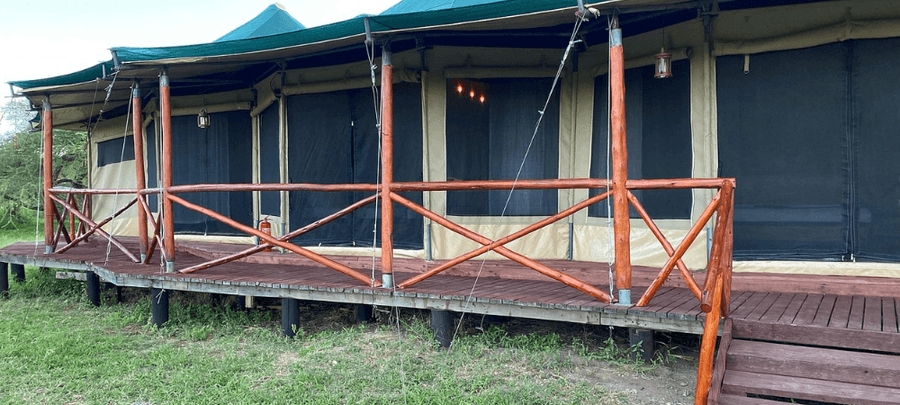 A spacious luxury safari tent stands on a raised wooden deck surrounded by grassy wilderness. The tent features screened entrances, canvas walls, and rustic wooden railings, blend…