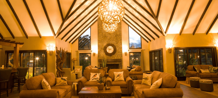 Warm and inviting lodge-style lounge with high vaulted ceilings, rustic wooden beams, glowing pendant lights, cozy seating areas, and a central stone fireplace creating an elegant…