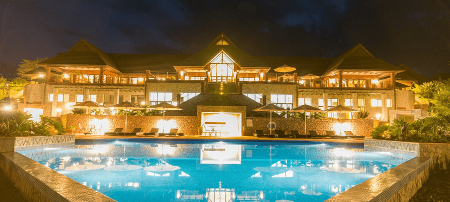 Luxurious resort illuminated at night, featuring a large glowing swimming pool, elegant terraces, warm ambient lighting, and dramatic architecture under a dark evening sky.