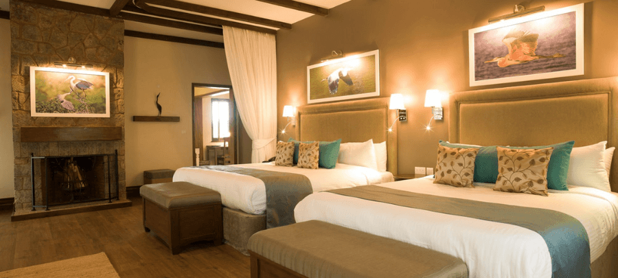 Spacious luxury hotel room with two large beds, elegant décor, warm lighting, wooden floors, and a cozy stone fireplace creating a welcoming atmosphere.