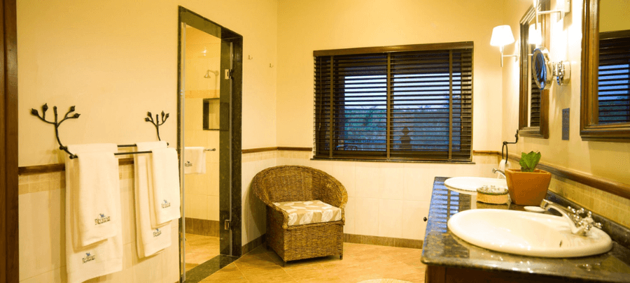 Spacious and elegant bathroom featuring double sinks, warm lighting, a glass-enclosed shower, wicker chair seating area, and large windows with wooden blinds.