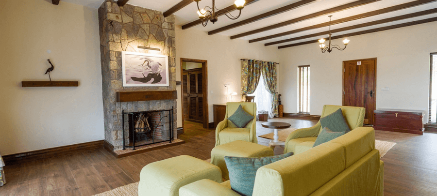 Spacious lodge-style living room with a stone fireplace, exposed wooden ceiling beams, comfortable green seating, wooden floors, and large windows with patterned curtains letting…