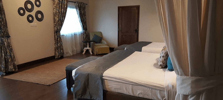 Warm and spacious bedroom featuring two canopy-style beds, dark wooden floors, patterned curtains, soft lighting, and a cozy seating area with an armchair and side table.
