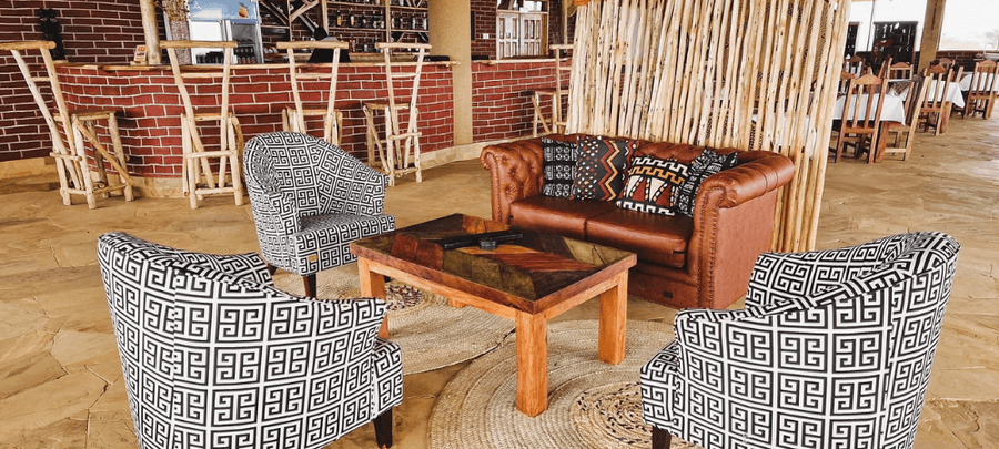 A stylish lounge seating area featuring patterned armchairs, a brown leather sofa with colorful cushions, and a wooden coffee table on woven rugs. In the background, a rustic bric…