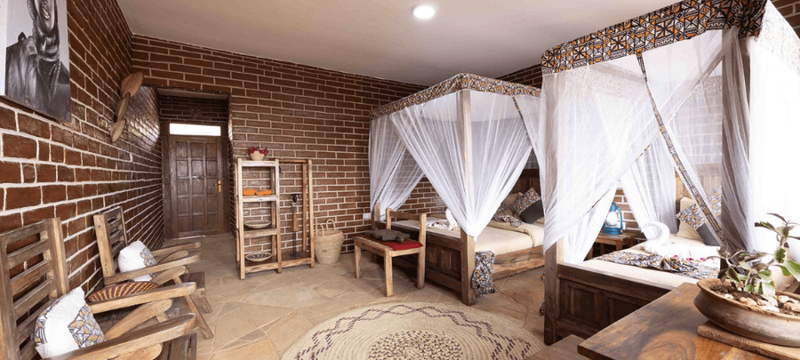A cozy rustic bedroom with exposed brick walls, wooden furniture, and a four-poster bed draped in white mosquito netting. The room features woven decor, cushioned wooden chairs, s…