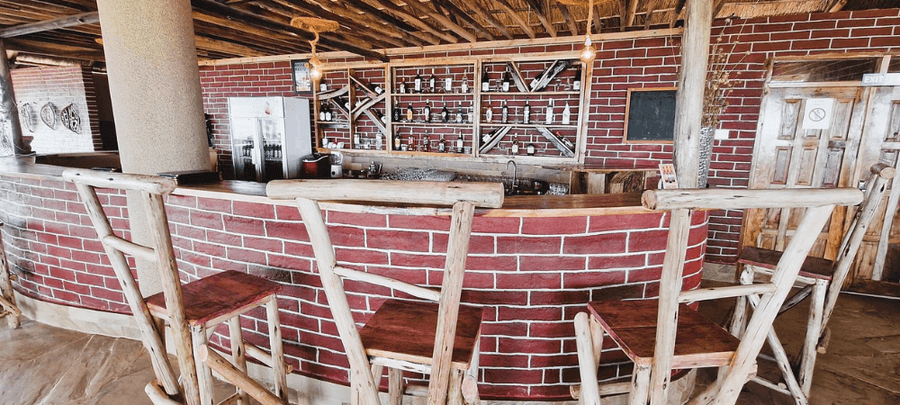 A rustic bar area with red brick walls, a curved wooden counter, and handcrafted wooden bar stools. Shelves stocked with bottles line the back wall beneath warm hanging lights, cr…