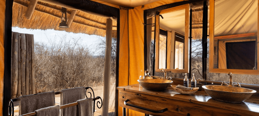 A safari-style bathroom with double basin sinks, wooden finishes, and large mirrors opens to an outdoor shower area overlooking dry bushland.