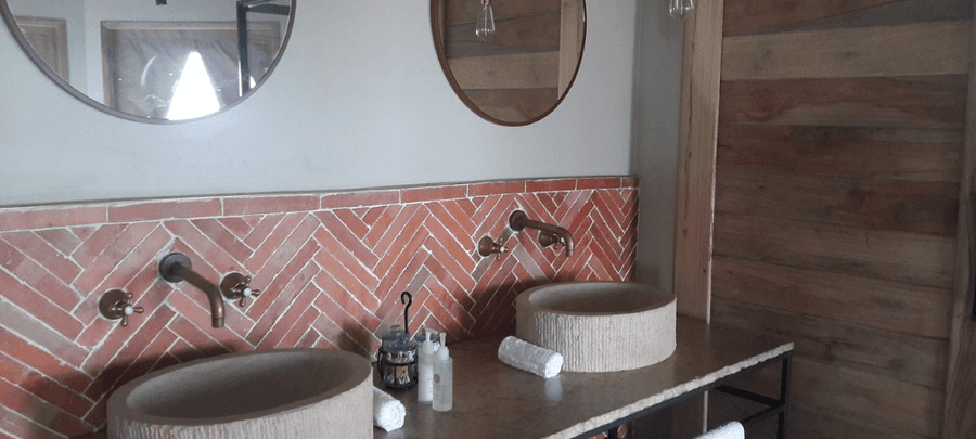 A stylish modern bathroom with two stone vessel sinks, round mirrors, brass wall-mounted faucets, and a red herringbone tile backsplash beside rustic wooden paneling.