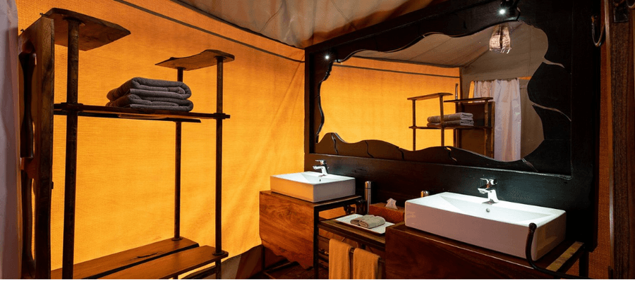 A stylish safari camp bathroom features dual modern sinks, warm ambient lighting, and dark wood finishes set against canvas tent walls. Open shelving with neatly folded towels and…