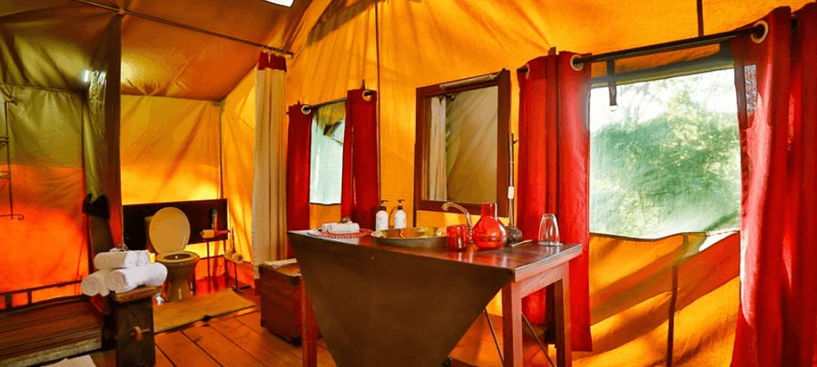 A warmly lit safari tent bathroom with canvas walls and red curtains features a wooden vanity with a basin sink, mirror, and decorative glass accents. The rustic interior also inc…
