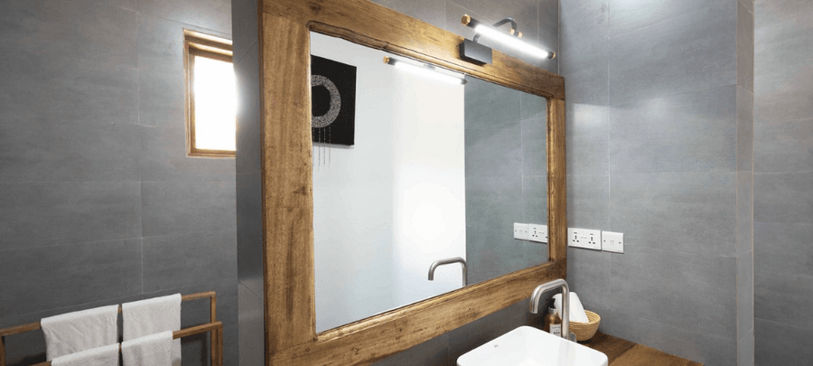 A modern bathroom interior with gray tiled walls, a large wooden-framed mirror, and a sleek white sink on a wooden countertop. Soft lighting, a small window, and neatly arranged t…