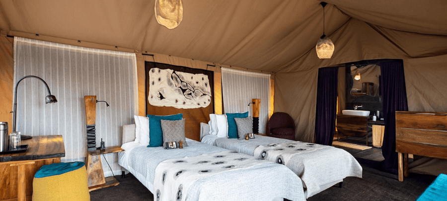 An elegant safari tent interior features two comfortable twin beds with soft linens, teal accent cushions, and handcrafted wooden furnishings. Warm lighting, canvas walls, and an…