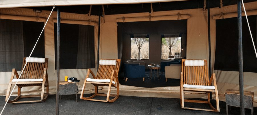 The entrance of a luxury safari tent features comfortable wooden rocking chairs and an open canvas front leading into a cozy lounge area. Soft natural lighting, modern seating, an…