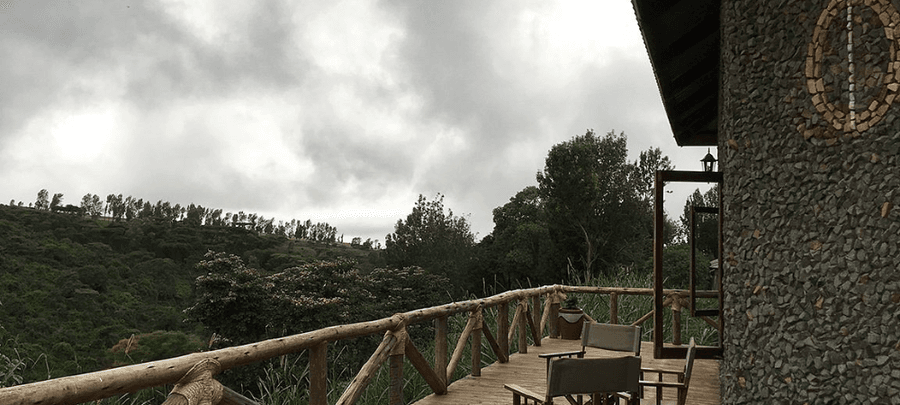 Rustic outdoor balcony with wooden railings and seating overlooking rolling green hills and dense forest under a cloudy sky.