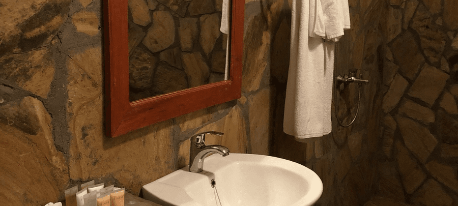 Rustic bathroom with natural stone walls, a white sink and mirror with wooden framing, hanging towels, and warm ambient lighting.