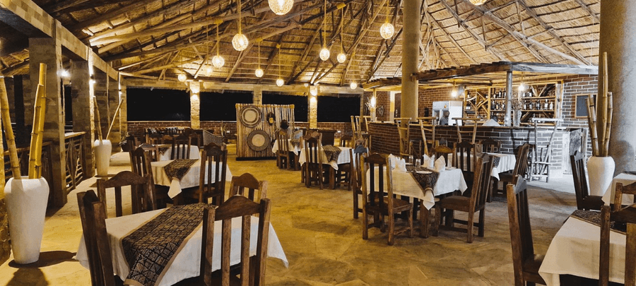 A spacious open-air restaurant with a rustic thatched roof, wooden dining tables, and warm hanging lights creating a cozy evening ambiance. The space includes a lodge-style bar ar…