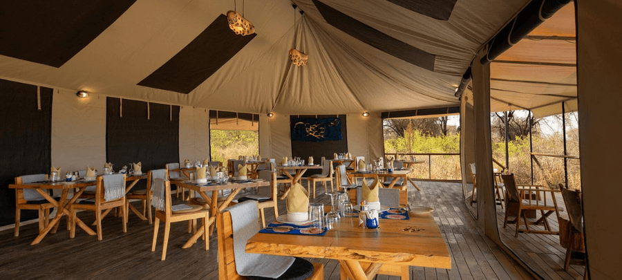 A spacious safari lodge dining area is set beneath a large canvas tent with wooden floors and open sides overlooking the surrounding bush. Elegantly arranged tables, rustic wooden…