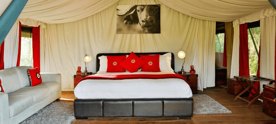 A stylish safari tent bedroom with white draped fabric walls and ceiling surrounds a large bed dressed in crisp white linens with bold red accent pillows and runners. Warm bedside…