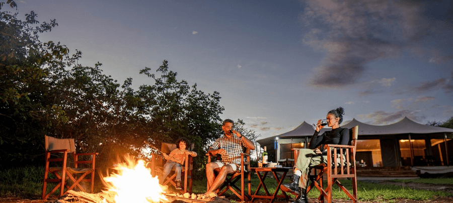 Guests relax around a glowing campfire at dusk outside a luxury safari tent camp, enjoying drinks and conversation under the open evening sky. Warm firelight contrasts with the so…