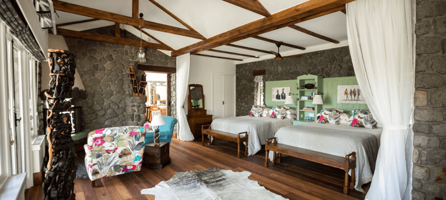 Spacious rustic-style bedroom with exposed wooden beams, stone walls, twin beds with colorful floral pillows, wooden flooring, and cozy seating near large windows.