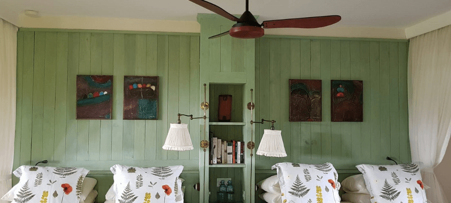 Cozy bedroom interior with a soft green paneled wall, decorative artwork, twin beds with patterned pillows, hanging bedside lamps, and a ceiling fan above.