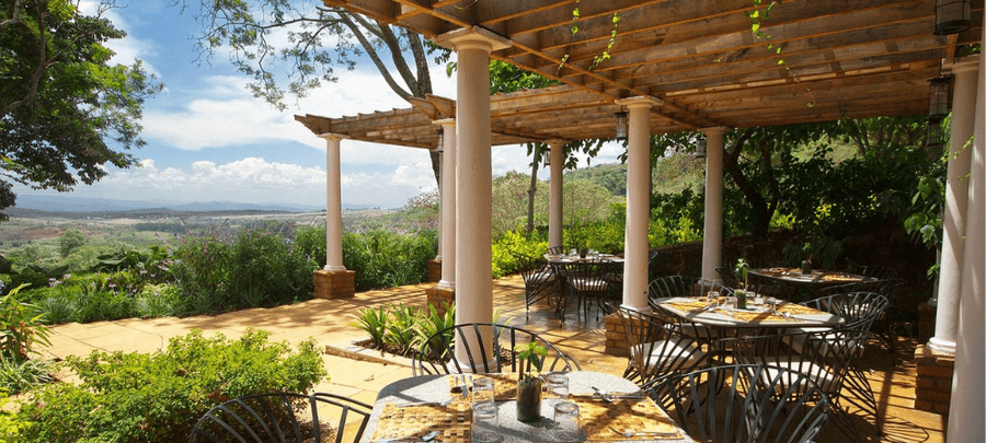 Scenic outdoor dining terrace with shaded pergolas, elegant table settings, lush gardens, and sweeping countryside views under a bright blue sky.